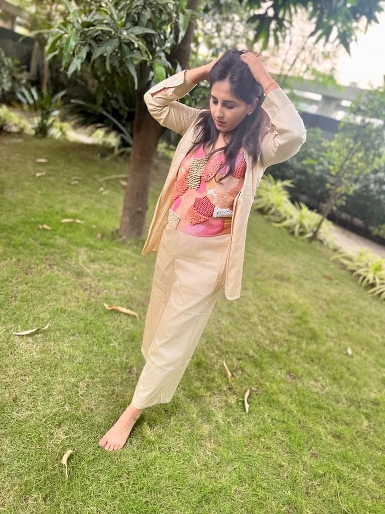 Woman in a garden wearing an open cream cotton blazer over a pink and coral beaded patchwork top with matching beige trousers.
